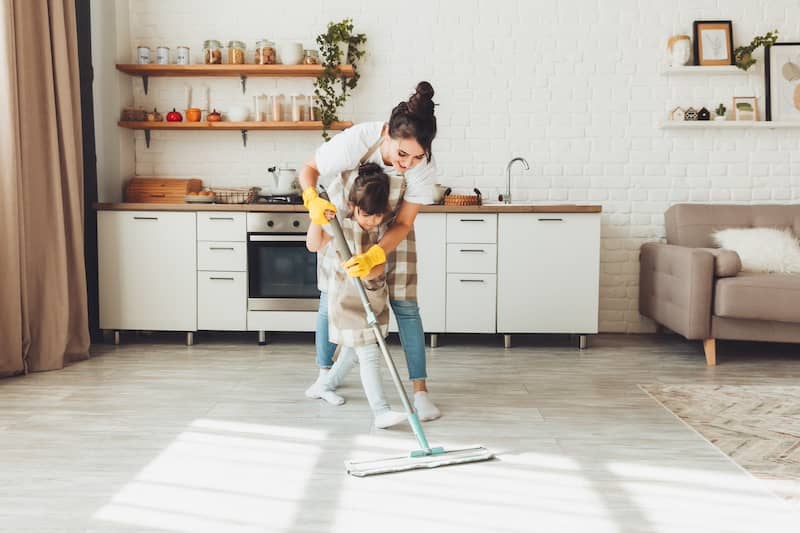 Mother and young daughter mop floor together in a well-organized kitchen.