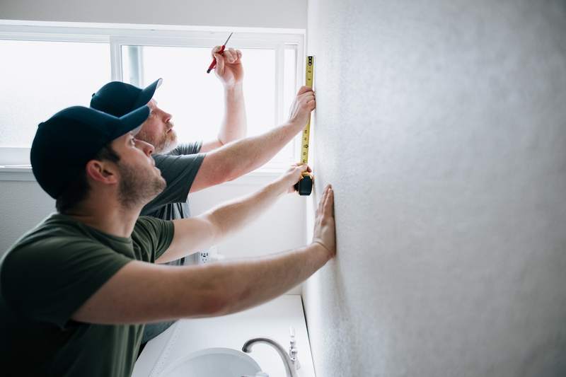Two men remodeling a bathroom hold a tape measure up against the wall to install a bathroom mirror above the sink.