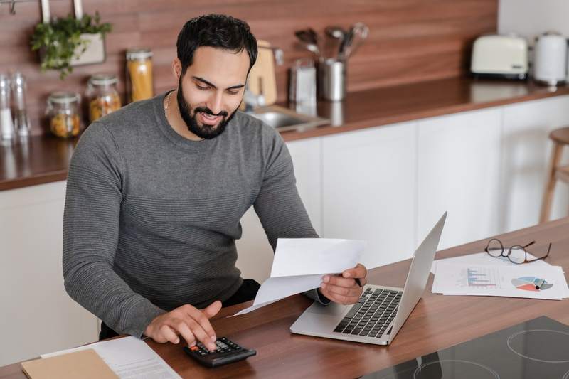 A man sits at his kitchen table with his laptop, holding a financial document and typing on a calculator.