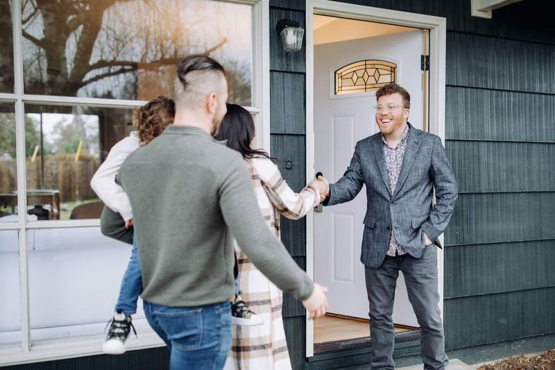 A real estate agent stands at the open door of a house and shakes hands with a woman whose husband and child are following behind her.