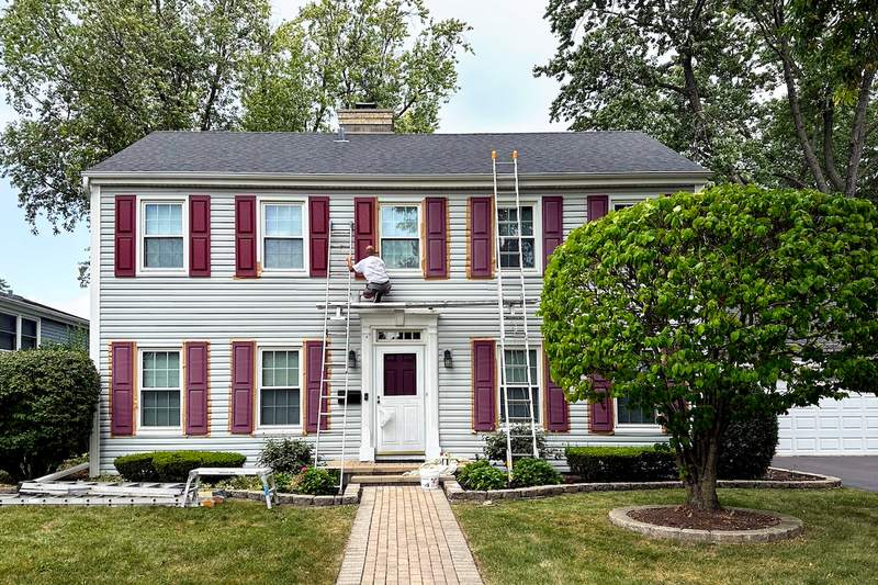 A man paints the shutters of a two-story house with a ladder leaning against it.