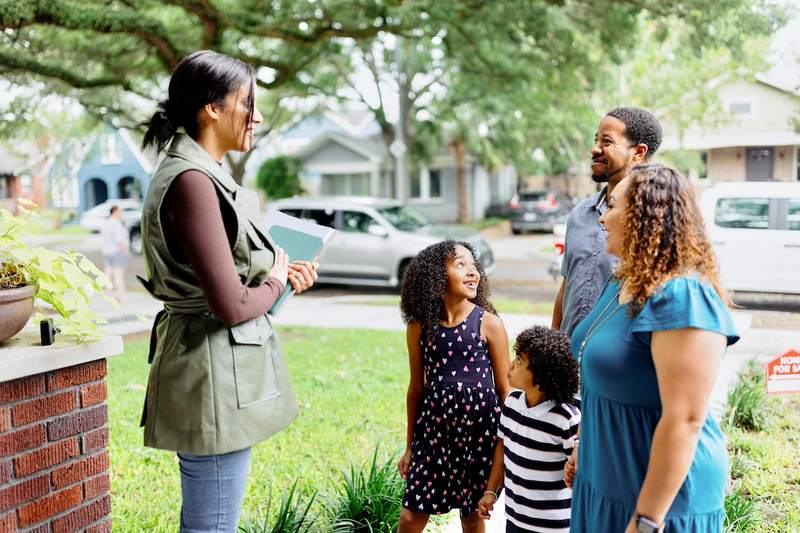 A real estate agent greets a family with two children on the front steps of a property for sale.